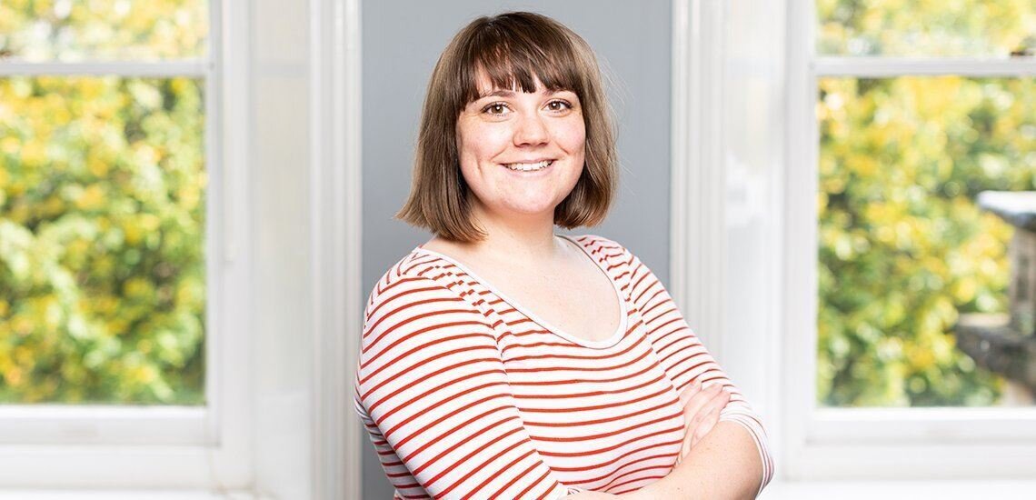 Caucasian woman smiles at the camera, she has a brunette bob hair cut and is wearing a white and red stripey top. She is stood in a spacious bright room with her arms crossed.